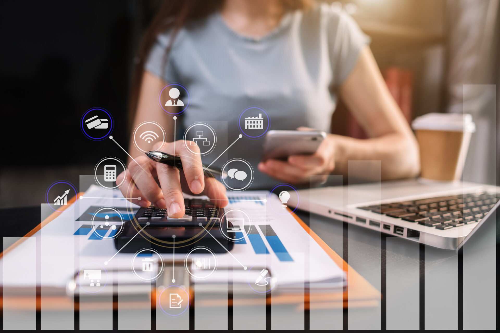 Zu sehen ist eine Frau an einem Schreibtisch. Vor ihr liegt ein Laptop und ein Taschenrechner, den sie mit einer Hand benutzt. In der anderen Hand bedient sie ein smartPhone.